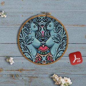 May include: A cross-stitch pattern featuring two blue figures with intricate floral designs, framed in a wooden hoop. The pattern is on a blue background and includes a PDF icon.