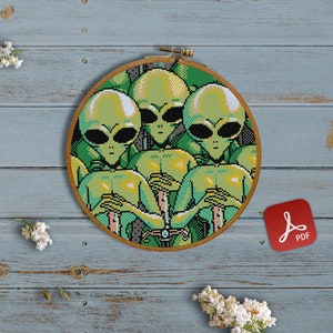 Pode incluir: Um padrão de ponto cruz de um grupo de alienígenas verdes com olhos grandes, sentados em fila. O padrão é emoldurado em um aro de madeira e está sobre um fundo de madeira azul.