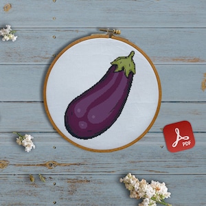 Puede incluir: Un patrón de punto de cruz de berenjena morada en un aro de bordado de madera. El aro está sobre una superficie de madera azul claro con flores blancas e hilo de bordar.