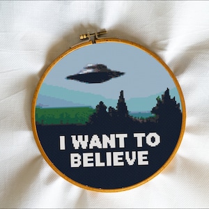 May include: A round, blue and white embroidery hoop with a pixelated image of a UFO flying over a forest and the text "I WANT TO BELIEVE".