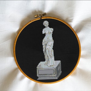 May include: A white cross-stitch embroidery of the Venus de Milo statue on a black background, framed in a wooden hoop.