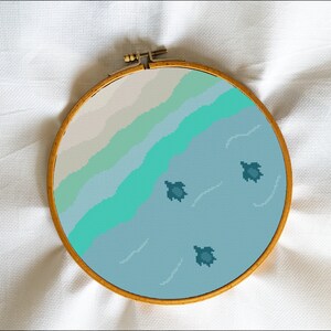 May include: A circular embroidery hoop featuring a pixelated design of a beach scene with three sea turtles swimming in the water, in shades of blue and green.