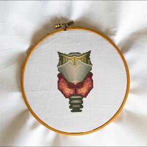 Puede incluir: Un aro de bordado de punto de cruz con una imagen pixelada de una criatura con un casco, un cuerpo rojo y naranja y una base verde y gris.