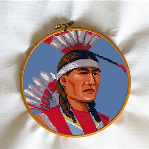 Puede incluir: Un bordado de punto de cruz que representa a un hombre nativo americano con un tocado de plumas. El hombre mira hacia la derecha y tiene una expresión seria en su rostro. El fondo es de color azul claro.