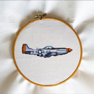 May include: A cross-stitch embroidery hoop with a pixelated image of a gray and orange airplane. The airplane has a white star on the wing and the number "08" on the side.
