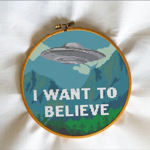 May include: A cross-stitch embroidery hoop with a blue and green landscape scene featuring a flying saucer and the text "I WANT TO BELIEVE".