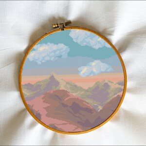 Op de afbeelding: Een borduurring met een kleurrijk landschapsscene. De scène toont een bergketen met een roze en paarse zonsondergangshemel en pluizige witte wolken.