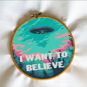 May include: A round embroidery hoop with a cross-stitch design of a UFO flying over a forest with the text "I WANT TO BELIEVE" in white.