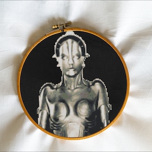 May include: A black and white cross-stitch embroidery of a robot-like figure with a metallic finish, framed in a wooden hoop.