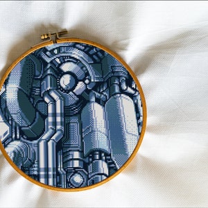 Può includere: Un design pixelato blu e grigio di un robot o una macchina è ricamato su un tessuto bianco in un cerchio da ricamo rotondo.