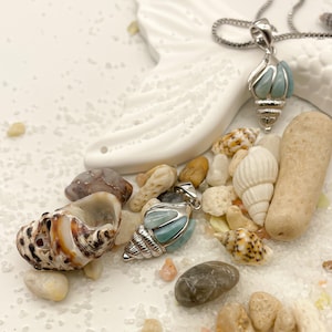 May include: Two silver shell-shaped pendants with blue and green gemstone accents, displayed on a white ceramic surface with seashells and pebbles.