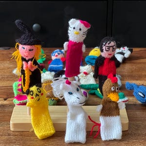 Op de afbeelding: Een set kleurrijke gebreide vingerpoppen, waaronder een heks, een eenhoorn, een kip, een Hello Kitty-personage en een blauwe vogel. De poppen worden getoond op een houten standaard.