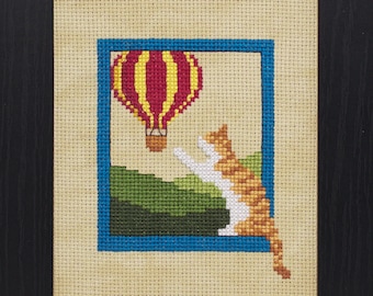 May include: Cross stitch pattern featuring a cat looking up at a hot air balloon in the sky. The pattern is framed in a black frame and is ready to be stitched.