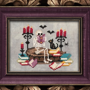 Peut inclure: Une œuvre d'art au point de croix dans un cadre violet avec le texte "Buried in Books". L'image représente un squelette lisant un livre, avec un chat noir, des bougies et des chauves-souris. L'œuvre est de Lola Crow Cross Stitch.
