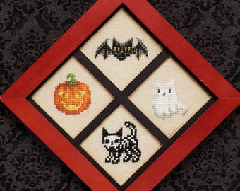 May include: A framed cross-stitch artwork featuring four Halloween-themed designs. The designs include a bat, a jack-o'-lantern, a ghost, and a skeleton cat. The frame is red, and the background is a dark, patterned fabric.