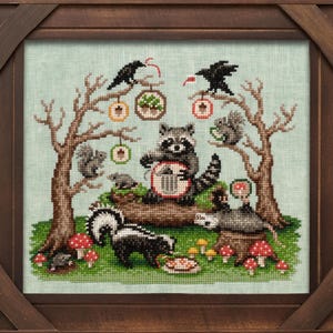May include: Cross-stitch artwork in a wooden frame. The scene depicts a raccoon playing a banjo, surrounded by squirrels, skunks, and mushrooms. Two black crows perch in the trees, with decorative ornaments hanging from the branches. The background is a light green.