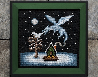 May include: A cross-stitch pattern featuring a white dragon blowing smoke onto a small green house in a snowy landscape. The dragon is flying in the night sky with a full moon in the background. The pattern is framed in a dark green frame with a black outer frame.