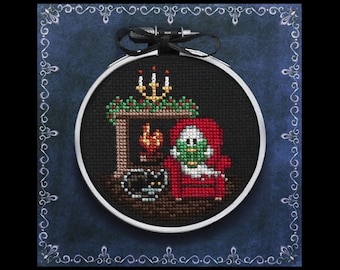 May include: A cross-stitch pattern for a Christmas ornament featuring a snowman sitting in a red armchair by a fireplace with a black cat. The ornament is in a round hoop with a black background.