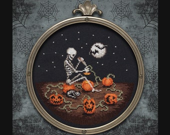 May include: A cross-stitch pattern depicting a skeleton sitting on a pumpkin, carving another pumpkin. The scene is set against a dark blue background with stars and a crescent moon. The pattern is framed in a gold-coloured hoop.