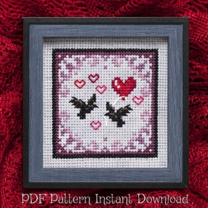 May include: A cross-stitch pattern featuring two bats flying with a red heart balloon and pink hearts on a purple and white background. The pattern is framed in a dark gray frame.