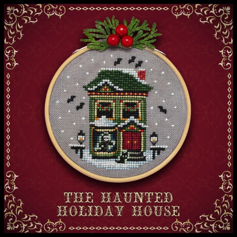 May include: A cross-stitch embroidery hoop with a gray background and a green house with a red door. The house has a snowy roof and is decorated with lights. The text "THE HAUNTED HOLIDAY HOUSE" is below the hoop.