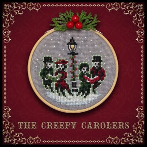 May include: A cross-stitch ornament featuring a group of skeletons singing carols under a street lamp. The ornament is framed in a wooden hoop and has a red background with gold accents. The text "THE CREEPY CAROLEERS" is printed below the ornament.