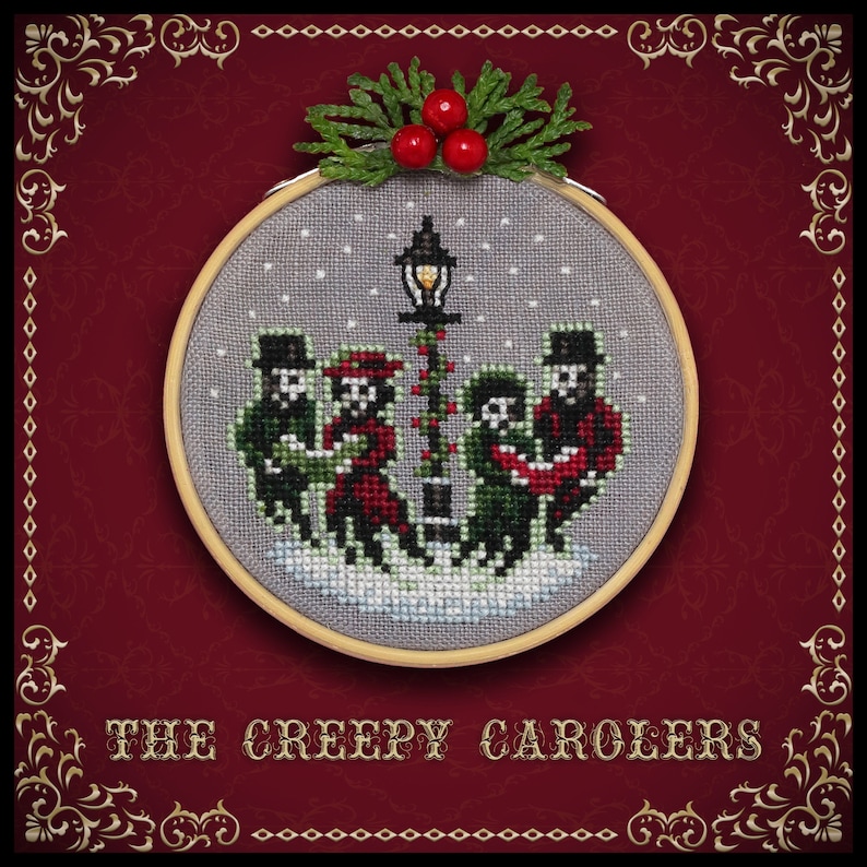 May include: A cross-stitch ornament featuring a group of skeletons singing carols under a street lamp. The ornament is framed in a wooden hoop and has a red background with gold accents. The text "THE CREEPY CAROLEERS" is printed below the ornament.