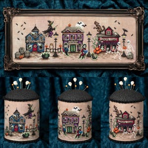 May include: A cross-stitch embroidery featuring a Halloween scene with a funeral parlor, library, and general store, displayed in a black ornate frame and on three cylindrical pin cushions topped with skull-headed pins on a dark fabric.