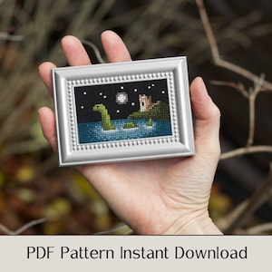 May include: A cross-stitch pattern depicting a green Loch Ness Monster swimming in a lake with a castle in the background. The pattern is framed in a silver frame and is held in a hand.