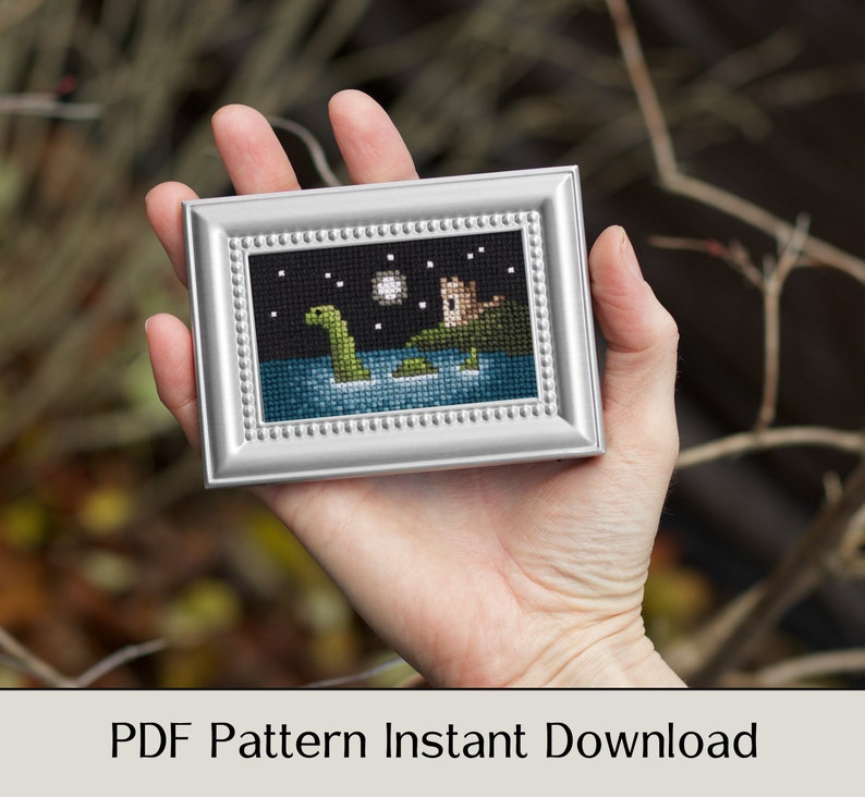 May include: A cross-stitch pattern depicting a green Loch Ness Monster swimming in a lake with a castle in the background. The pattern is framed in a silver frame and is held in a hand.