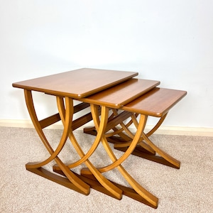 May include: A set of three wooden nesting tables. Each table has a rectangular top and curved, arch-shaped legs. The tables are finished in a medium-brown colour. The largest table is approximately 61 cm long.
