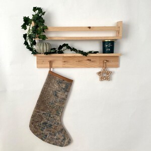 May include: A wooden shelf with two shelves and two hooks. The top shelf has a green candle and a small white planter with green vines. The bottom shelf has a brown stocking hanging from one of the hooks and a wooden star ornament hanging from the other hook.
