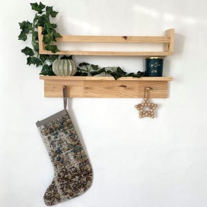 Puede incluir: Una estantería de madera con dos niveles y un gancho. La estantería está decorada con una planta verde, un pequeño jarrón de cerámica, una vela turquesa y un adorno de estrella de madera. Una media estampada cuelga del gancho.