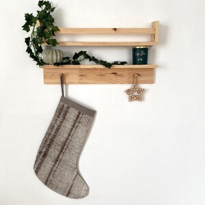 Op de afbeelding: Een houten plank met twee niveaus en een haak. Een grijze en bruine sok hangt aan de haak. Een klein houten sterretje hangt aan de onderste plank. Een groene kaars pot staat op de bovenste plank.