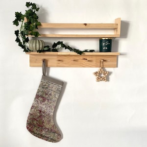 May include: A wooden shelf with two shelves and hooks. The top shelf has a green candle and a small gray planter with a green plant. The bottom shelf has a stocking with a colorful pattern and a wooden star ornament.