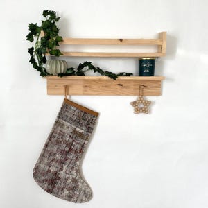 Puede incluir: Una estantería de madera con dos estantes y un gancho. Un tarro de vela verde está en el estante superior. Una media con un puño de cuero marrón cuelga del gancho. Un adorno de estrella de madera cuelga del estante inferior.