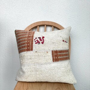May include: Cream-colored decorative pillow with a patchwork design. The pillow features rectangular and square patches in shades of cream, brown, and red. The pillow is placed on a wooden chair.
