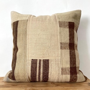 May include: A square decorative pillow with a patchwork design. The pillow features a mix of beige and brown fabrics, including stripes and plaid patterns. The pillow is placed on a wooden surface.