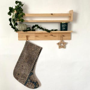 Puede incluir: Una estantería de madera con dos estantes y ganchos, una vela verde, un pequeño jarrón de cerámica con enredaderas verdes y una media marrón y azul colgando de un gancho. Un adorno de estrella de madera cuelga de otro gancho.