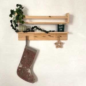 May include: A wooden shelf with two tiers and a hook. The shelf has a green candle, a small gray planter with a green plant, and a red and white stocking hanging from the hook. A wooden star ornament hangs from the bottom shelf.