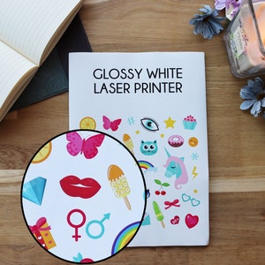 May include: A close-up of a glossy white laser printer with a sticker sheet featuring colorful cartoon images. The sticker sheet has a variety of images including a butterfly, a unicorn, a cat, a donut, a cupcake, a popsicle, a rainbow, a heart, a star, and a lightning bolt. The text "Glossy White Laser Printer" is printed on the top of the printer.