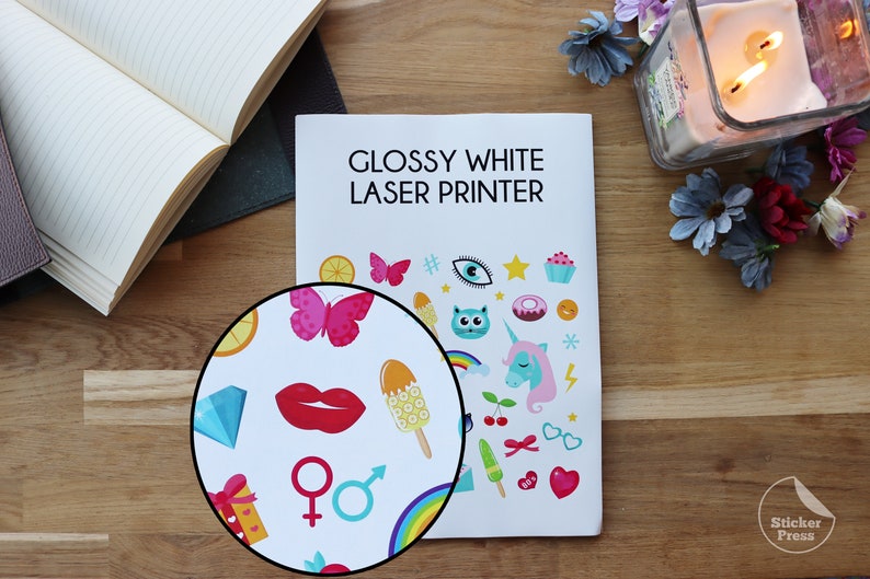 May include: A close-up of a glossy white laser printer with a sticker sheet featuring colorful cartoon images. The sticker sheet has a variety of images including a butterfly, a unicorn, a cat, a donut, a cupcake, a popsicle, a rainbow, a heart, a star, and a lightning bolt. The text "Glossy White Laser Printer" is printed on the top of the printer.