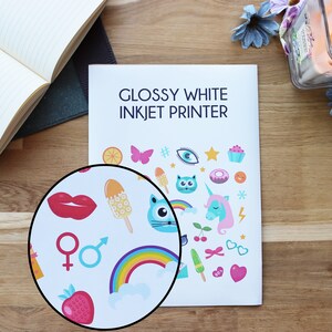 May include: Glossy white inkjet printer paper with a sheet of colorful stickers printed on it. The stickers feature a variety of fun and whimsical designs, including a rainbow, a unicorn, a cat, a cupcake, and a strawberry.