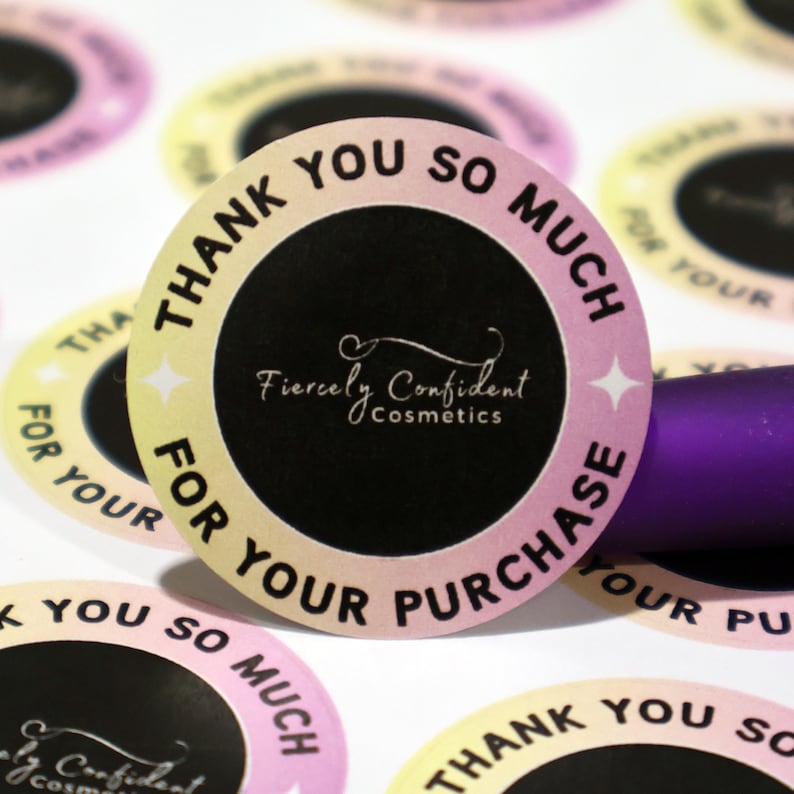 Puede incluir: Pegatina redonda de color rosa, amarillo y negro con el texto "Muchas gracias por su compra" y el logotipo "Fiercely Confident Cosmetics".