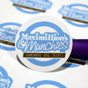 Puede incluir: Una pegatina redonda blanca con un c&iacute;rculo azul y una pancarta marr&oacute;n. La pegatina dice "Maximillion's Munchies Homemade Dog Treats" en texto blanco con un gr&aacute;fico de huella de pata.
