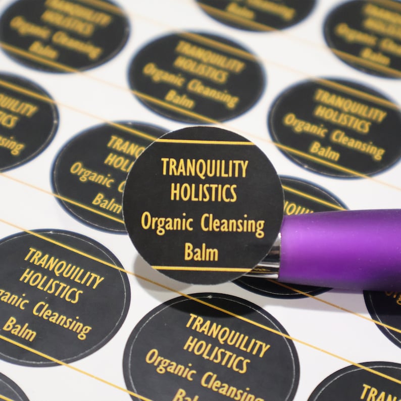 Puede incluir: Primer plano de una pegatina redonda negra con letras doradas que dice "Tranquility Holistics Organic Cleansing Balm".