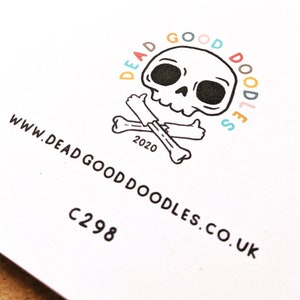 May include: A white card with a black and white illustration of a skull and crossbones. The skull is smiling and giving a thumbs up. The text "Dead Good Doodles" is written in a circle around the skull, with each letter in a different color. The text "www.deadgooddoodles.co.uk" is written below the skull and crossbones. The text "C298" is written below the website address.