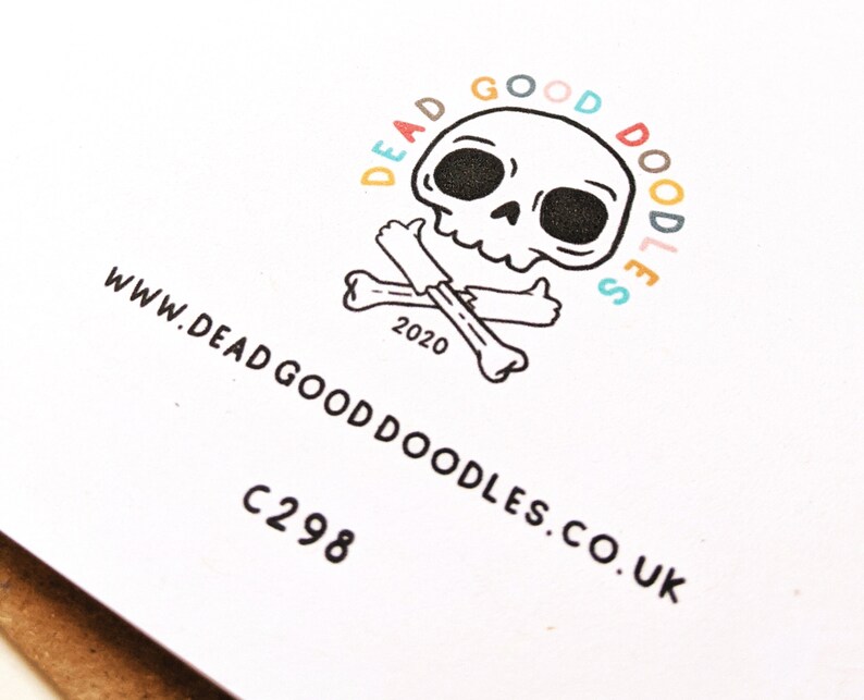 May include: A white card with a black and white illustration of a skull and crossbones. The skull is smiling and giving a thumbs up. The text "Dead Good Doodles" is written in a circle around the skull, with each letter in a different colour. The text "www.deadgooddoodles.co.uk" is written below the skull and crossbones. The text "C298" is written below the website address.