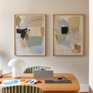 May include: Two framed abstract paintings in neutral tones of beige, grey, and black. The artwork features layered shapes and lines. A white mushroom lamp, striped box, and desk accessories are on a wooden desk.