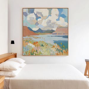 May include: A framed landscape painting depicting a tranquil scene. The artwork shows a blue sky with fluffy white clouds, a body of water, and colourful land with mountains in the background. The painting is in a wooden frame.